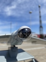 A close-up view of a drone or surveillance equipment mounted on a stand outdoors. The lens or sensor is encased in a transparent dome, with a clear blue sky and blurred structures in the background.