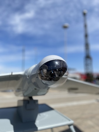 A close-up view of a drone or surveillance equipment mounted on a stand outdoors. The lens or sensor is encased in a transparent dome, with a clear blue sky and blurred structures in the background.