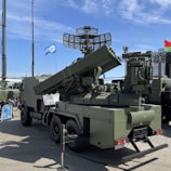 A military vehicle equipped with large and complex radar systems and missile launchers is displayed outdoors on a paved surface. It is painted in a camouflage green color and features a mounted rotating radar antenna. Several flags are visible in the background, indicating a possible exhibition or display setting.
