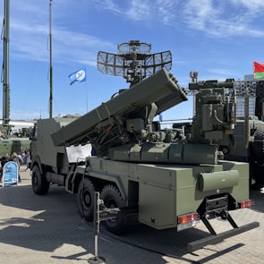 A military vehicle equipped with large and complex radar systems and missile launchers is displayed outdoors on a paved surface. It is painted in a camouflage green color and features a mounted rotating radar antenna. Several flags are visible in the background, indicating a possible exhibition or display setting.