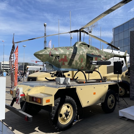 A military-style drone with camouflage paint is mounted on a platform with wheels, suggesting mobility for transport. The drone features helicopter-like rotors and visible equipment or attachments. Nearby are flags and people, possibly indicating an exhibition or event setting, with a clear blue sky in the background.