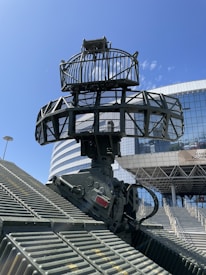 A large radar system is mounted on a diagonal grid-like structure with a modern building in the background. The radar is composed of cylindrical and circular components, mostly metallic in appearance, and is situated under a clear blue sky. The building displays reflective, glass paneling and is accompanied by signage that suggests an event or exhibition.