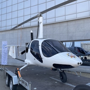 A white gyroplane is displayed on a trailer, positioned outdoors in front of a large, grid-patterned building. The gyroplane features a sleek design, with its rotor blades positioned above the body and a distinct star emblem on the front. Reflections of buildings are visible in the dark, tinted windows of the gyroplane.