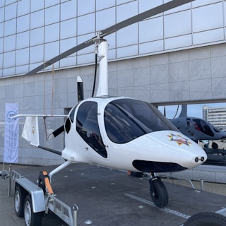 A white gyroplane is displayed on a trailer, positioned outdoors in front of a large, grid-patterned building. The gyroplane features a sleek design, with its rotor blades positioned above the body and a distinct star emblem on the front. Reflections of buildings are visible in the dark, tinted windows of the gyroplane.
