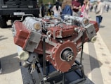A large, industrial engine with a metallic and red-hued exterior is mounted on a black stand. It features multiple pistons, cylinders, and components, suggesting a complex mechanical design. In the background, several people are gathered in casual attire, standing on a paved surface.