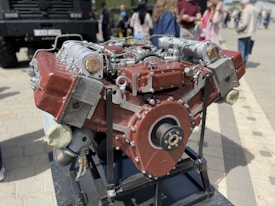 A large, industrial engine with a metallic and red-hued exterior is mounted on a black stand. It features multiple pistons, cylinders, and components, suggesting a complex mechanical design. In the background, several people are gathered in casual attire, standing on a paved surface.