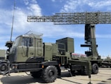 A military radar system mounted on a large, green military vehicle. The radar's structure is prominently displayed above the vehicle, showcasing its intricate metal framework. The vehicle has durable, large wheels suitable for rough terrains. The sky is clear with a few clouds, suggesting it is daytime. The surrounding area is paved, indicating a display or vehicle exhibition.