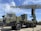 A military radar system mounted on a large, green military vehicle. The radar's structure is prominently displayed above the vehicle, showcasing its intricate metal framework. The vehicle has durable, large wheels suitable for rough terrains. The sky is clear with a few clouds, suggesting it is daytime. The surrounding area is paved, indicating a display or vehicle exhibition.