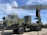 A military radar system mounted on a large, green military vehicle. The radar's structure is prominently displayed above the vehicle, showcasing its intricate metal framework. The vehicle has durable, large wheels suitable for rough terrains. The sky is clear with a few clouds, suggesting it is daytime. The surrounding area is paved, indicating a display or vehicle exhibition.
