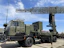 A military radar system mounted on a large, green military vehicle. The radar's structure is prominently displayed above the vehicle, showcasing its intricate metal framework. The vehicle has durable, large wheels suitable for rough terrains. The sky is clear with a few clouds, suggesting it is daytime. The surrounding area is paved, indicating a display or vehicle exhibition.