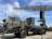 A military radar system mounted on a large, green military vehicle. The radar's structure is prominently displayed above the vehicle, showcasing its intricate metal framework. The vehicle has durable, large wheels suitable for rough terrains. The sky is clear with a few clouds, suggesting it is daytime. The surrounding area is paved, indicating a display or vehicle exhibition.