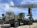 A military radar system mounted on a large, green military vehicle. The radar's structure is prominently displayed above the vehicle, showcasing its intricate metal framework. The vehicle has durable, large wheels suitable for rough terrains. The sky is clear with a few clouds, suggesting it is daytime. The surrounding area is paved, indicating a display or vehicle exhibition.