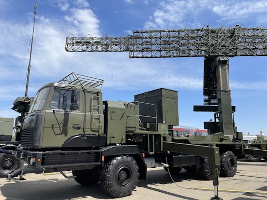 A military radar system mounted on a large, green military vehicle. The radar's structure is prominently displayed above the vehicle, showcasing its intricate metal framework. The vehicle has durable, large wheels suitable for rough terrains. The sky is clear with a few clouds, suggesting it is daytime. The surrounding area is paved, indicating a display or vehicle exhibition.