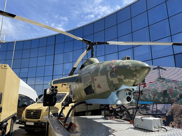 A helicopter with a camouflage design on display in an outdoor setting. It is positioned on a platform surrounded by vehicles and against the backdrop of a modern glass building reflecting the sky.