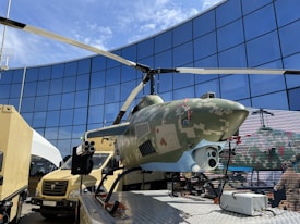 A helicopter with a camouflage design on display in an outdoor setting. It is positioned on a platform surrounded by vehicles and against the backdrop of a modern glass building reflecting the sky.