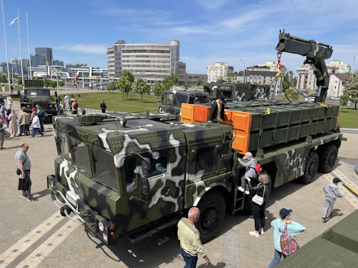A military truck with camouflage pattern is parked outdoors, surrounded by a group of people observing it. The truck is equipped with orange containers and a crane attached at the back. In the background, modern buildings and a green park area can be seen.