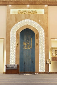The interior space features an ornate archway with intricate Islamic geometric patterns and Arabic calligraphy. A blue panel with additional calligraphy is situated at the center, flanked by two decorative columns. On the floor, a pair of microphones is set up, indicating this area might be used for speeches or prayers. A cushioned seat is placed on the left, and a music stand with a book is on the right.