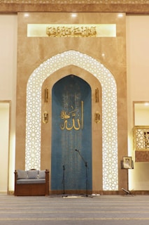 The interior space features an ornate archway with intricate Islamic geometric patterns and Arabic calligraphy. A blue panel with additional calligraphy is situated at the center, flanked by two decorative columns. On the floor, a pair of microphones is set up, indicating this area might be used for speeches or prayers. A cushioned seat is placed on the left, and a music stand with a book is on the right.