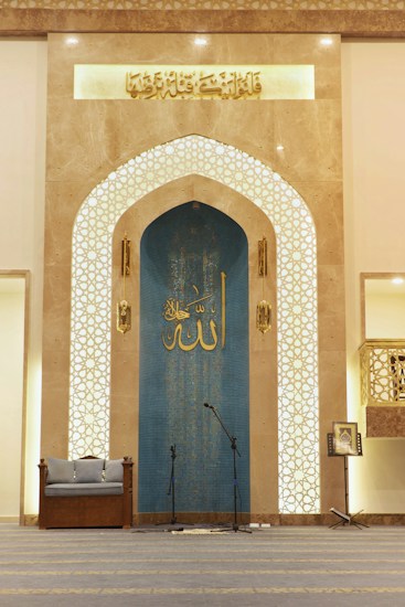 The interior space features an ornate archway with intricate Islamic geometric patterns and Arabic calligraphy. A blue panel with additional calligraphy is situated at the center, flanked by two decorative columns. On the floor, a pair of microphones is set up, indicating this area might be used for speeches or prayers. A cushioned seat is placed on the left, and a music stand with a book is on the right.