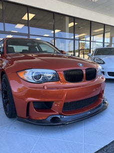 An orange high-performance car is parked on a tiled floor inside a glass-fronted showroom. A white car is visible in the background through the windows, reflecting the interior lighting.