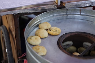 A traditional tandoor oven with round pieces of dough being baked on its surface. The oven appears to be part of a cart, suggesting a street food setup. The dough pieces are golden brown and some appear to have green herbs embedded in them.