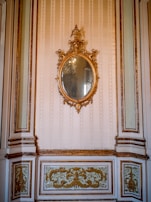 An ornate, oval-shaped mirror with an intricate gilded frame hangs on a wall. The wall features elegant molding and decorative panels with gold accents. The overall design is sophisticated and opulent, suggestive of a historical or classical interior.