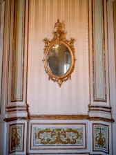 An ornate, oval-shaped mirror with an intricate gilded frame hangs on a wall. The wall features elegant molding and decorative panels with gold accents. The overall design is sophisticated and opulent, suggestive of a historical or classical interior.