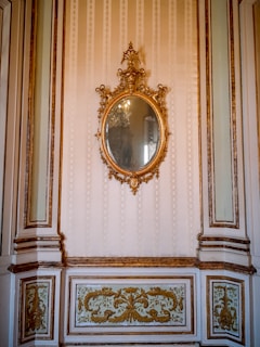 An ornate, oval-shaped mirror with an intricate gilded frame hangs on a wall. The wall features elegant molding and decorative panels with gold accents. The overall design is sophisticated and opulent, suggestive of a historical or classical interior.