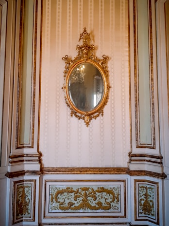 An ornate, oval-shaped mirror with an intricate gilded frame hangs on a wall. The wall features elegant molding and decorative panels with gold accents. The overall design is sophisticated and opulent, suggestive of a historical or classical interior.