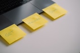 Three yellow sticky notes are attached to the edge of a laptop, each with a word written on them forming the phrase 'GET THINGS DONE'. The laptop is partially visible and the notes are placed on a white surface.