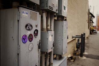 A row of utility boxes is mounted on an exterior brick wall in an urban alleyway. The boxes are adorned with various stickers including logos and designs, and some graffiti is visible. The background shows a weathered wall, a narrow passage, and blurred urban elements.