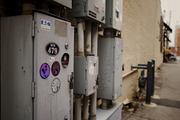 A row of utility boxes is mounted on an exterior brick wall in an urban alleyway. The boxes are adorned with various stickers including logos and designs, and some graffiti is visible. The background shows a weathered wall, a narrow passage, and blurred urban elements.