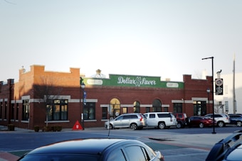 A street view of a red brick building with a green sign that reads 'Dollar Saver'. Several cars are parked in front of the building, and there are street lamps lining the sidewalk. The sky is clear and slightly overcast, suggesting late afternoon.