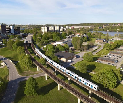 Aerial view of a light rail or metro train traveling on an elevated track through a suburban area with lush greenery and residential buildings. The scene includes roads and a body of water in the background under a clear blue sky.
