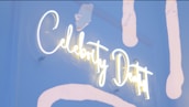 A neon sign with the words 'Celebrity Dentist' is displayed. The sign is lit with bright white and yellow neon lights, set against a background with light blue and white abstract paint strokes. Some lights appear slightly dim at the edges.