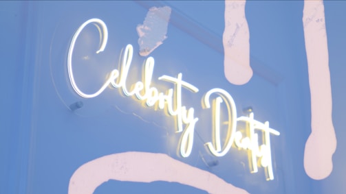 A neon sign with the words 'Celebrity Dentist' is displayed. The sign is lit with bright white and yellow neon lights, set against a background with light blue and white abstract paint strokes. Some lights appear slightly dim at the edges.