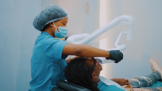 A dental professional wearing blue scrubs, a mask, and a hair cover is performing a procedure on a patient lying in a dental chair. The professional is holding a piece of dental equipment over the patient's mouth. The setting is likely a dental clinic with muted blue and white colors.