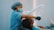 A dental professional wearing blue scrubs, a mask, and a hair cover is performing a procedure on a patient lying in a dental chair. The professional is holding a piece of dental equipment over the patient's mouth. The setting is likely a dental clinic with muted blue and white colors.