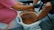 A person in pink scrubs assists another person with a foot bath. The person providing the service wears black gloves, and the other person's feet are submerged in a light blue basin filled with brownish water. The setting suggests a pedicure or spa treatment.