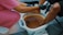 A person in pink scrubs assists another person with a foot bath. The person providing the service wears black gloves, and the other person's feet are submerged in a light blue basin filled with brownish water. The setting suggests a pedicure or spa treatment.