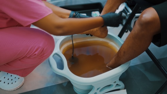 A person in pink scrubs assists another person with a foot bath. The person providing the service wears black gloves, and the other person's feet are submerged in a light blue basin filled with brownish water. The setting suggests a pedicure or spa treatment.