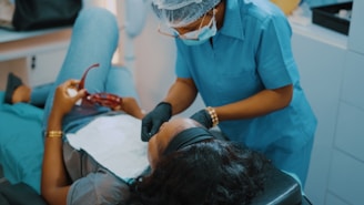 A dental professional wearing a blue uniform and protective gear leans over a patient who is reclining in a dental chair. The patient holds a pair of red glasses in their hand, and a dental bib is placed over their torso.