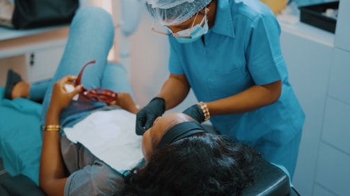 A dental professional wearing a blue uniform and protective gear leans over a patient who is reclining in a dental chair. The patient holds a pair of red glasses in their hand, and a dental bib is placed over their torso.