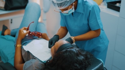 A dental professional wearing a blue uniform and protective gear leans over a patient who is reclining in a dental chair. The patient holds a pair of red glasses in their hand, and a dental bib is placed over their torso.