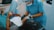 A dental professional wearing a blue uniform and protective gear leans over a patient who is reclining in a dental chair. The patient holds a pair of red glasses in their hand, and a dental bib is placed over their torso.
