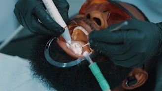 A close-up view of a dental procedure taking place, showing a patient lying with their mouth open. The patient is wearing protective eyewear, and two hands wearing black gloves are holding dental instruments inside the patient's mouth.