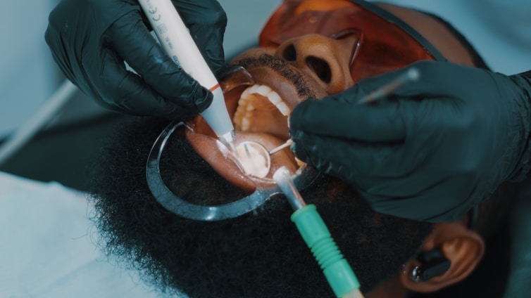 A close-up view of a dental procedure taking place, showing a patient lying with their mouth open. The patient is wearing protective eyewear, and two hands wearing black gloves are holding dental instruments inside the patient's mouth.