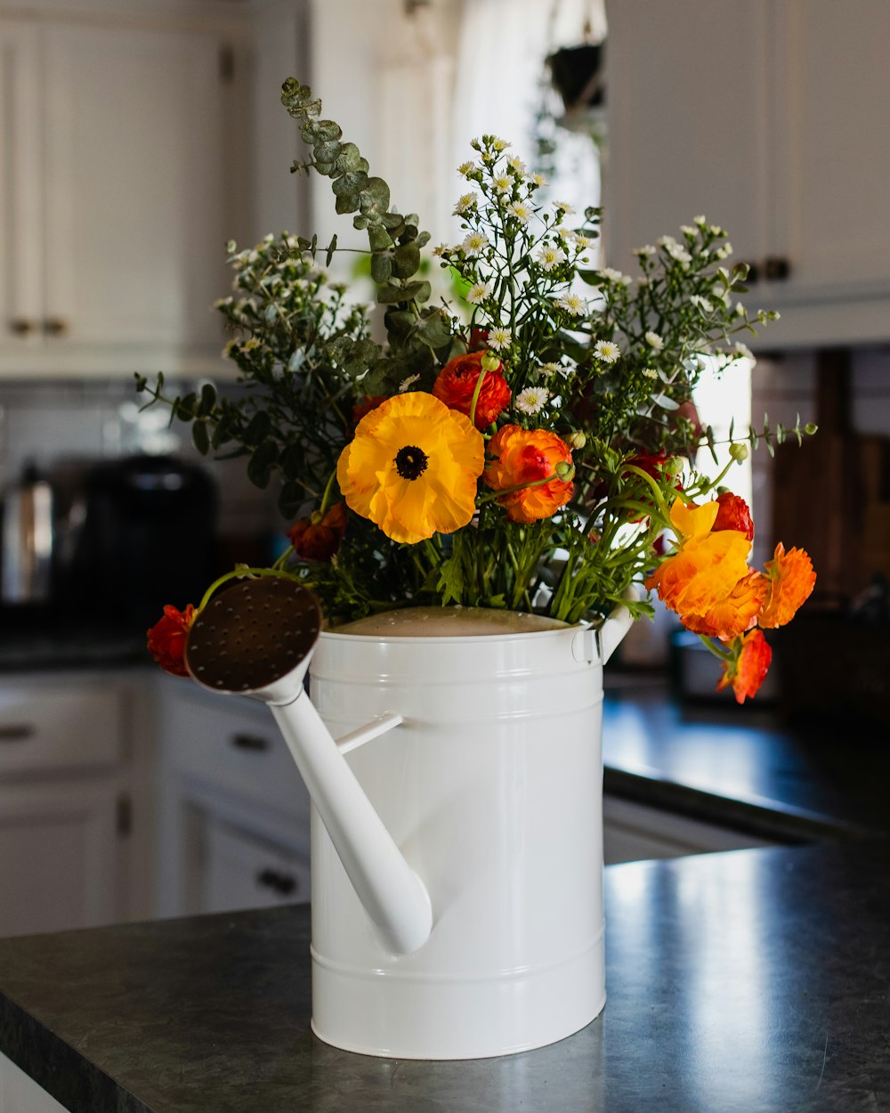 a white watering can filled with flowers on a kitchen counter