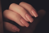A close-up view of a hand with long, sparkly pink glitter nail polish on the fingernails. The nails are neatly manicured and have a glossy finish, emphasizing the sparkles in the polish.