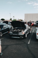 Wide shot of the car club gathering with members inspecting different vehicles under bright daylight.